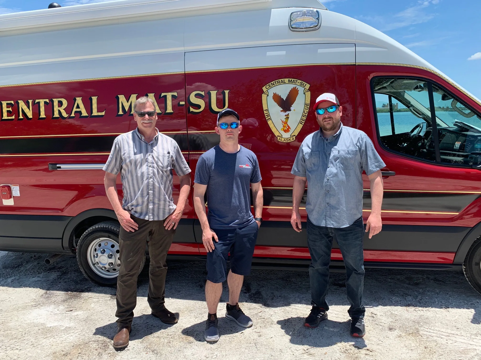 Three individuals standing beside a Frontline Communications CRU-22 Mobile Command vehicle on a sunny day.