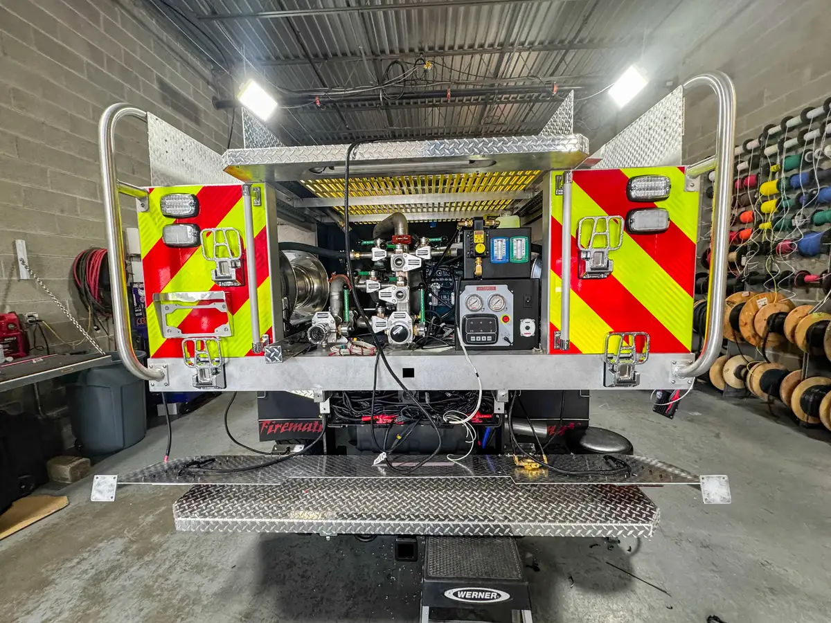 Rear-left view showing open rear doors, pump controls, hose reel side bay, and step platform.