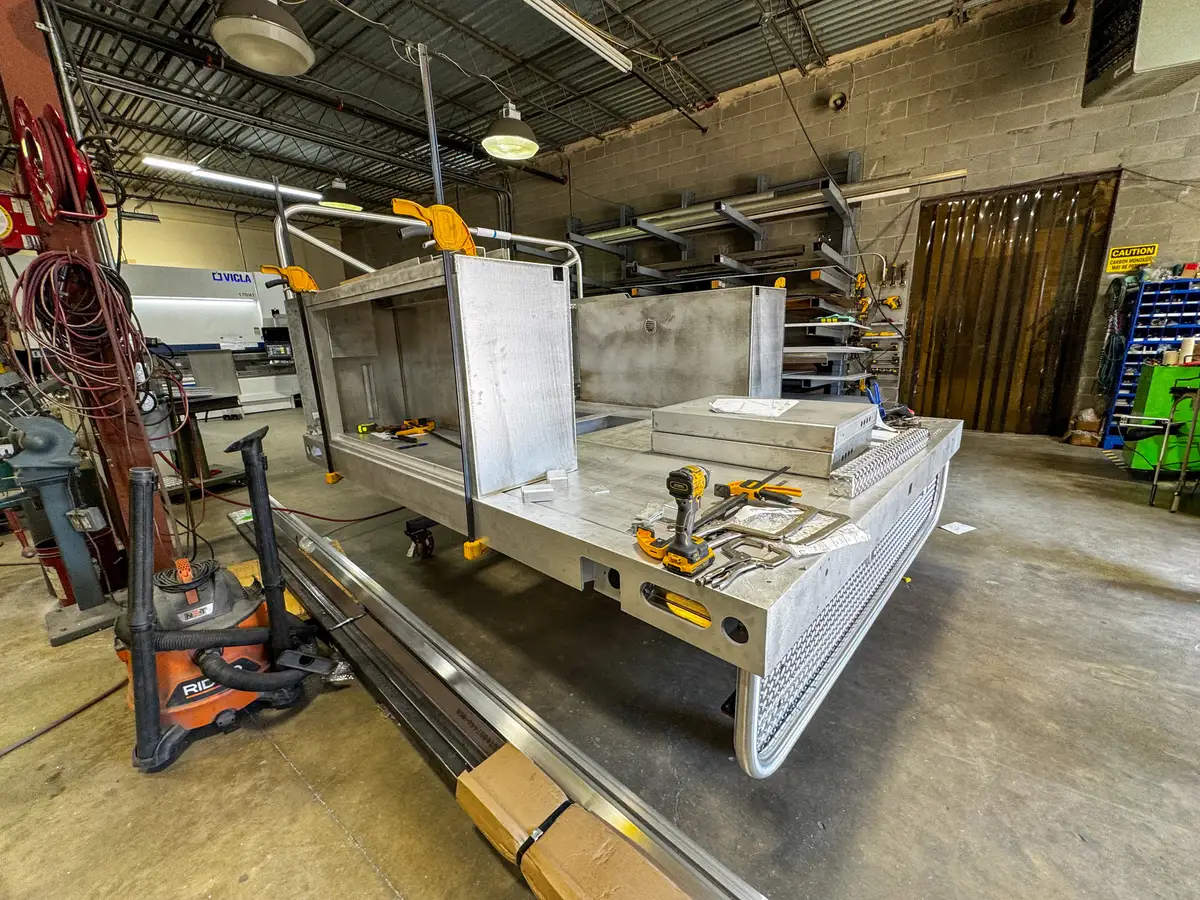 Rear-left view of the unfinished body module showing open deck area, compartment walls, and shop tools.