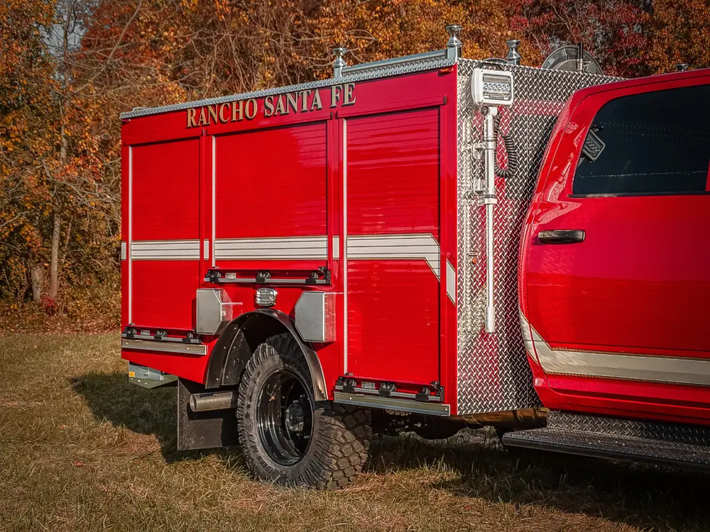 Exterior view of small fire truck showing cab, body compartments, and wheel/tire area.