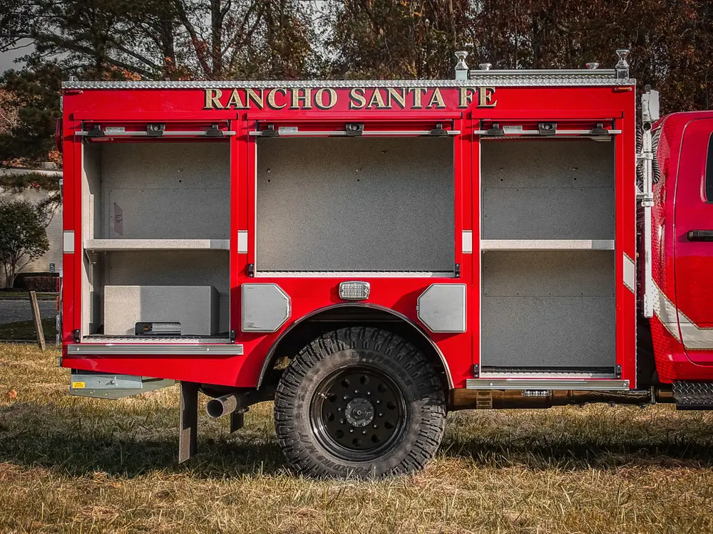 Exterior view of small fire truck showing cab, body compartments, and wheel/tire area.