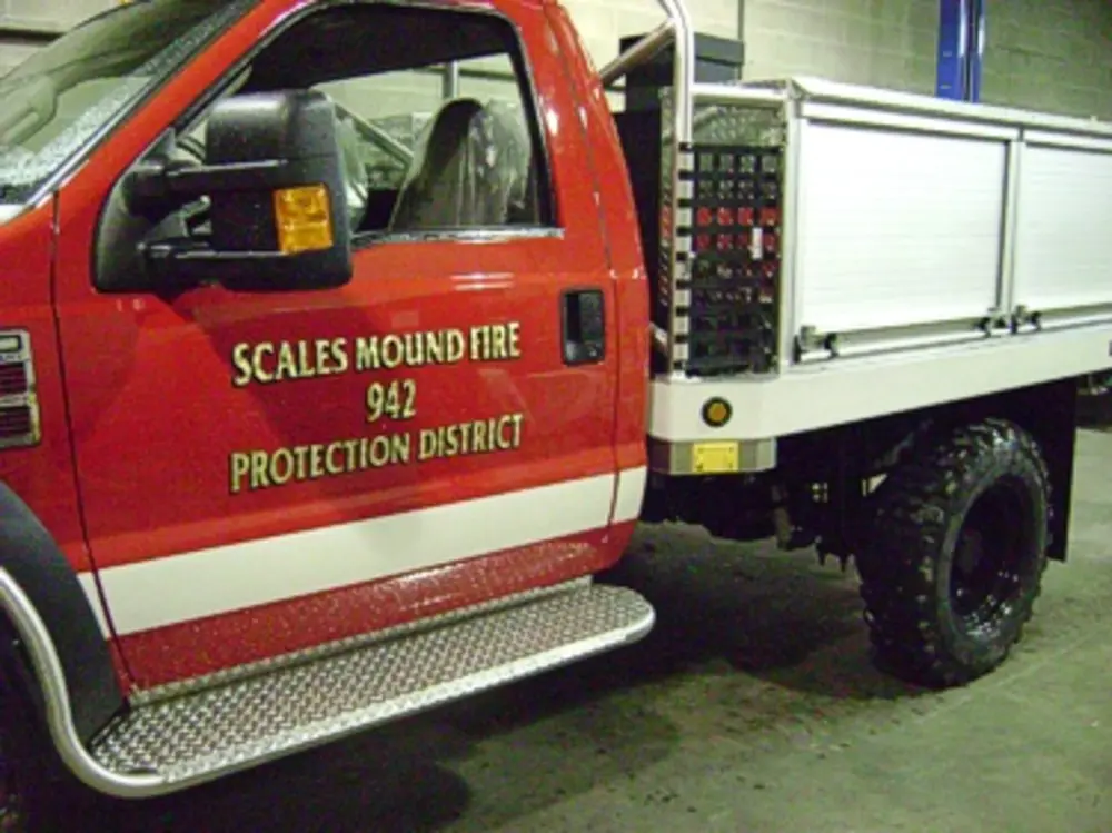 Exterior view of small fire truck showing cab, body compartments, and wheel/tire area.