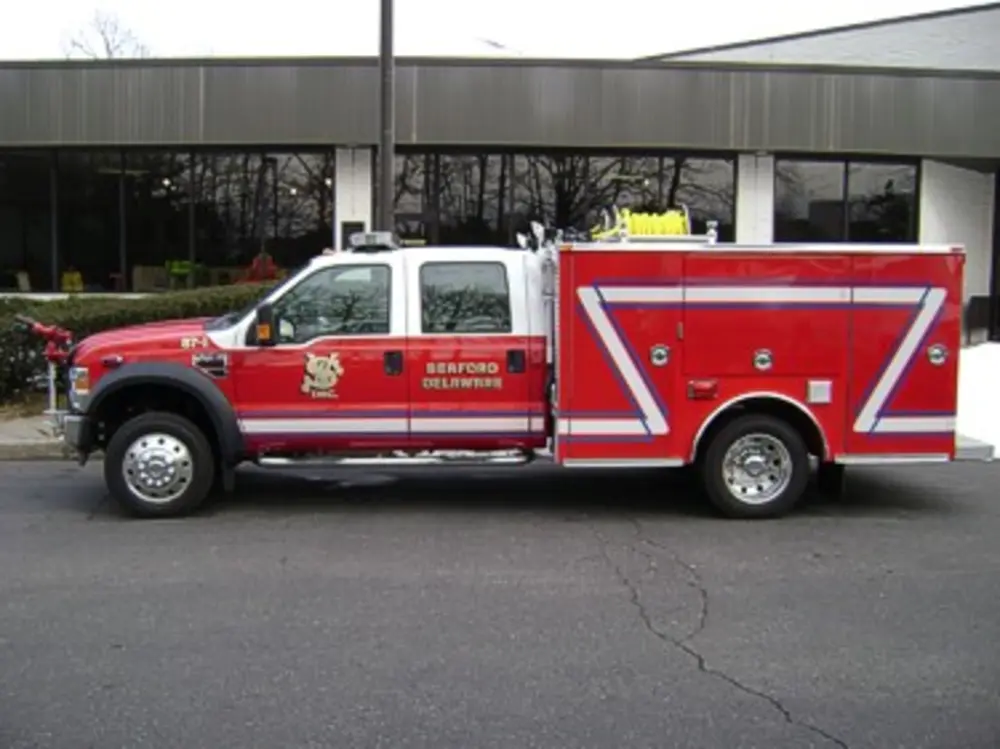 Exterior view of small fire truck showing cab, body compartments, and wheel/tire area.