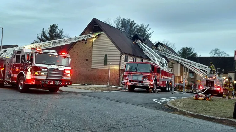 Fireground street view with two aerial apparatus, one ladder extended to a roofline and emergency lighting active.
