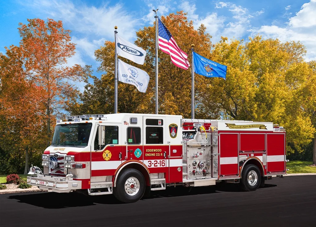 Front-left profile of a pumper beneath flagpoles showing cab face, pump panel section, and rear body.