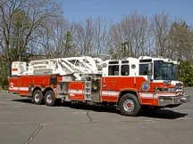 Wide front right view of the parked tower ladder with cab, stowed aerial, and rear body compartments.