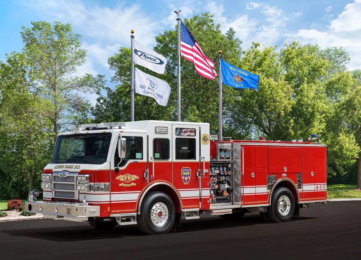 Front-left profile view with the pumper parked beneath flagpoles showing full side body and wheelbase.
