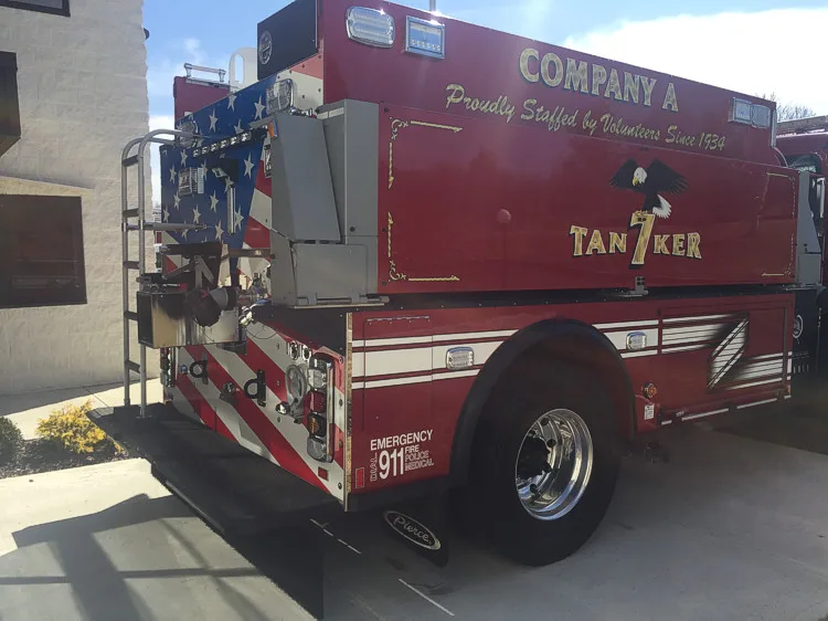 Rear left side view showing tank body graphics, rear discharge panel, wheel area, and tailboard.