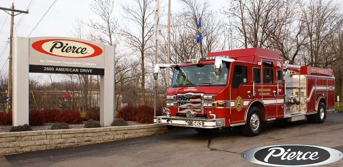 Front-left wide view of the pumper at the Pierce sign with side pump panel section visible.