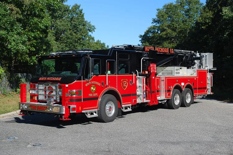 Front-left wide exterior view of the tower ladder with stowed boom, side compartments, and tandem rear wheels.