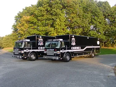 Wide side view of two parked black hazmat trucks with matching graphics and roof warning lights.