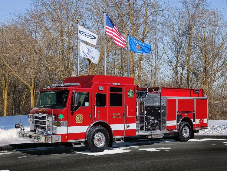 Front-left exterior view of a pumper with clover emblem showing cab, pump panel, and side compartments.